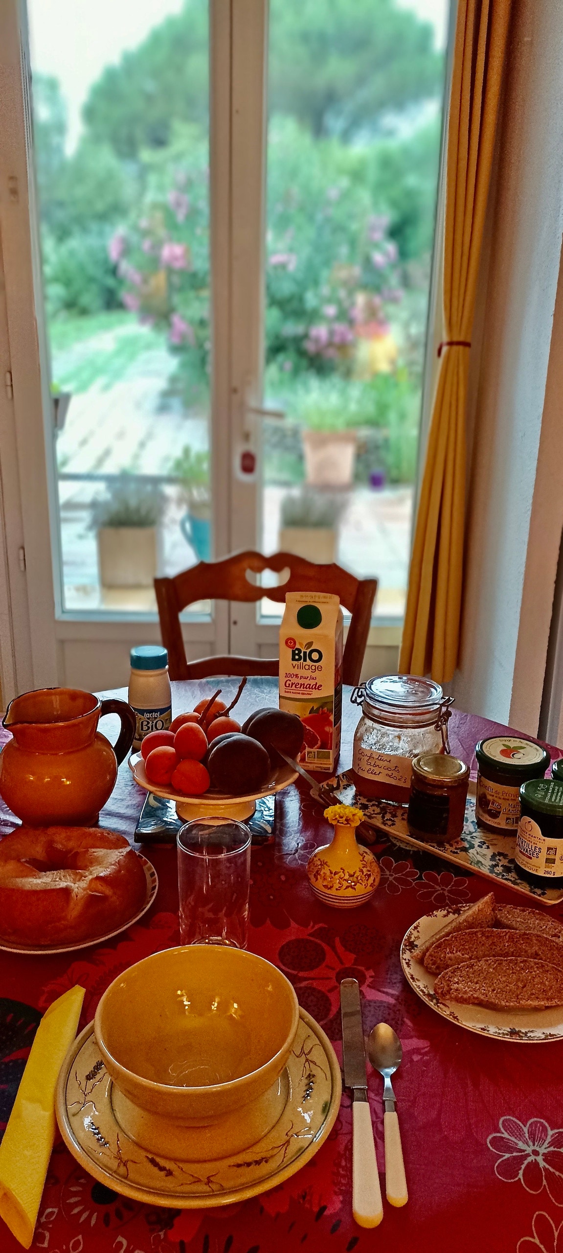 A dining table is set with a bright tablecloth, featuring bowls, jars, and various breakfast items. Fresh fruit, bread, and beverage containers are arranged, while a window framed by yellow curtains offers a view of a lush garden.