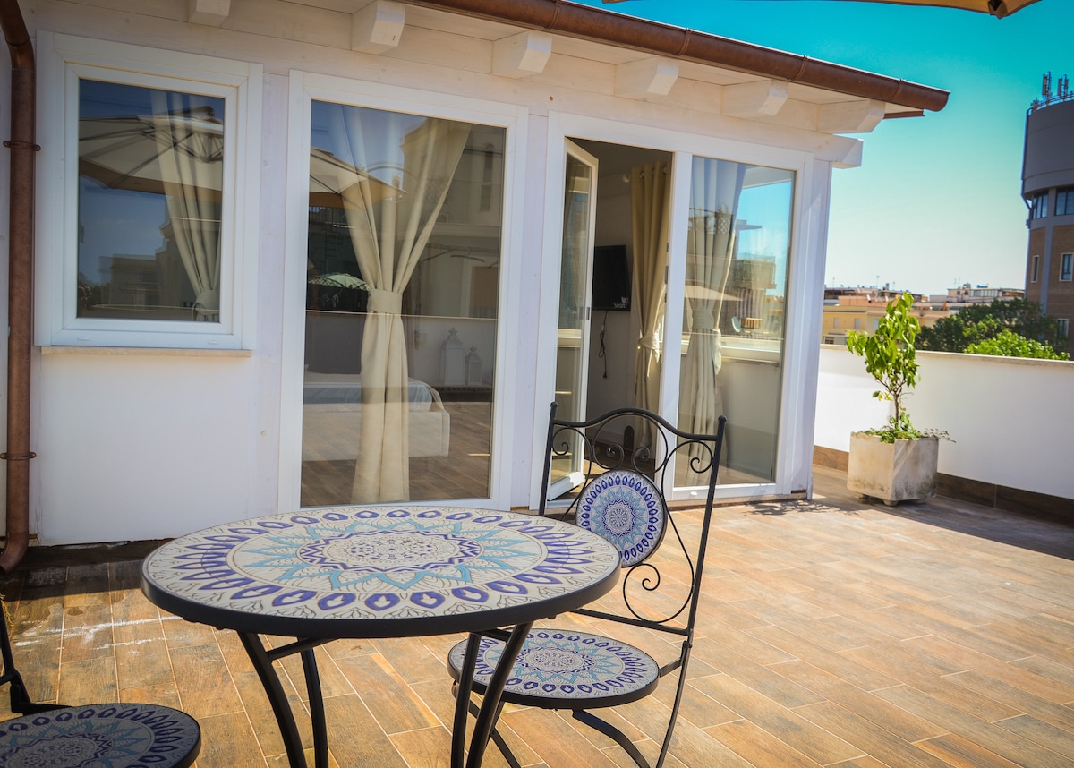 A sunlit terrace features a round table with intricately patterned tiles, accompanied by two matching chairs. Large glass doors provide access to an interior space, while light curtains frame the entrance, creating a seamless transition between indoor and outdoor areas.
