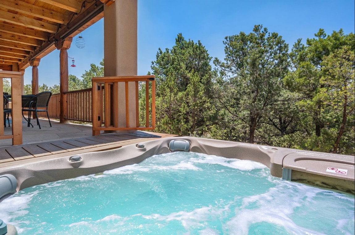 A hot tub filled with bubbling water is positioned on a wooden deck, surrounded by lush greenery. A sunlit porch can be seen in the background, offering seating for outdoor relaxation.