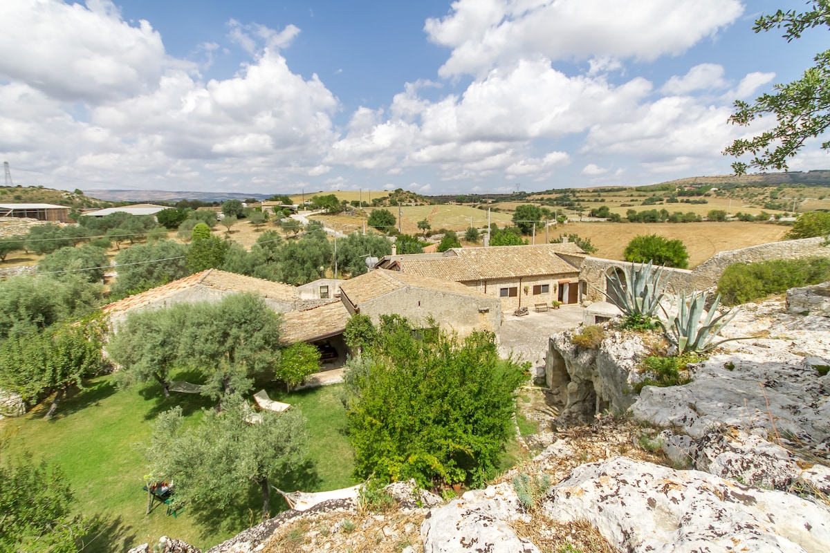Aerial view of Santolìo Country Retreat highlights the rustic architecture surrounded by lush greenery. Olive trees and shrubs populate the expansive garden, while the sloping hills in the background frame the serene countryside landscape under a partly cloudy sky.