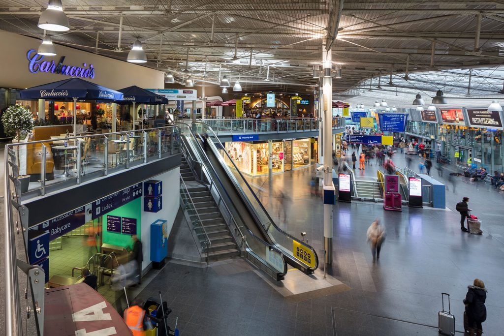 The interior of a large transportation hub is shown, featuring multiple levels and an escalator. Various shops and dining options are visible, with travelers moving throughout the space. Natural light filters in from a skylight above, illuminating the bustling atmosphere.