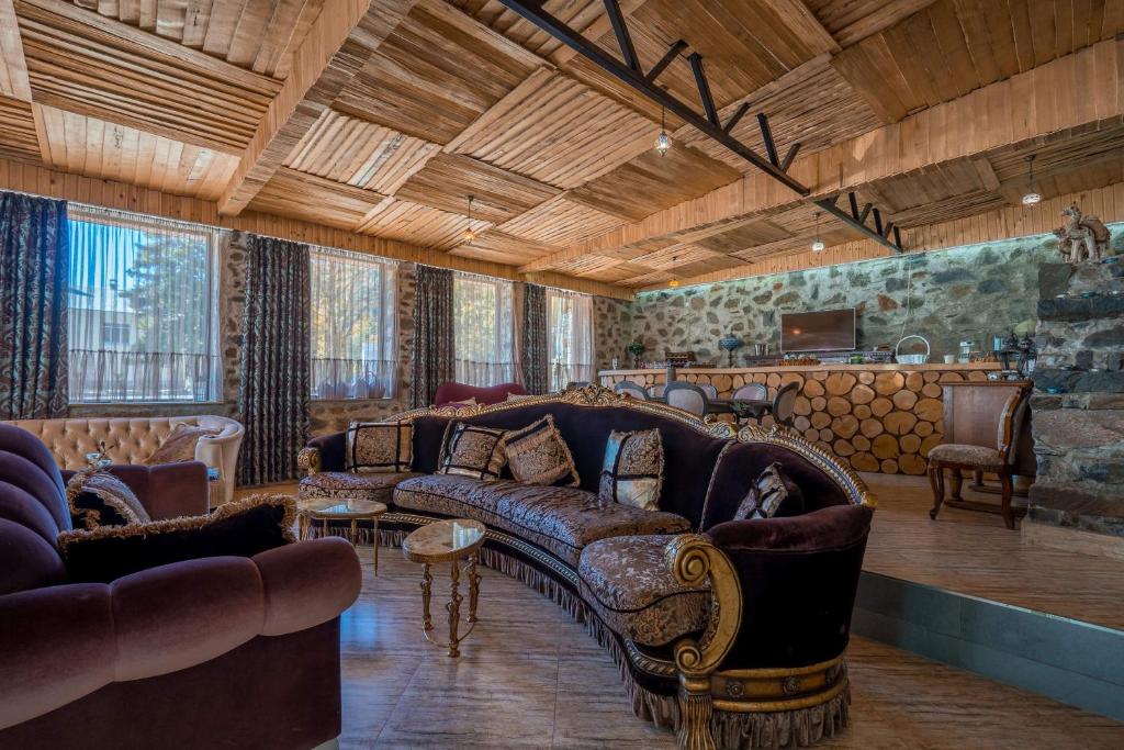 An inviting living area features a large curved sofa with rich upholstery, surrounded by natural wood ceilings and stone walls. Large windows permit ample light, and the open layout connects to a modern kitchen area adorned with wooden accents.