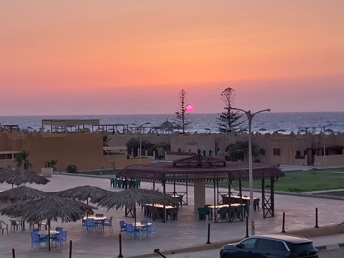 A serene coastal view captures the sunset as the sun meets the horizon, casting a soft pink hue over the sea. Umbrella-covered tables and chairs are arranged in the foreground, while structures dot the shoreline, all set against a backdrop of gentle waves.