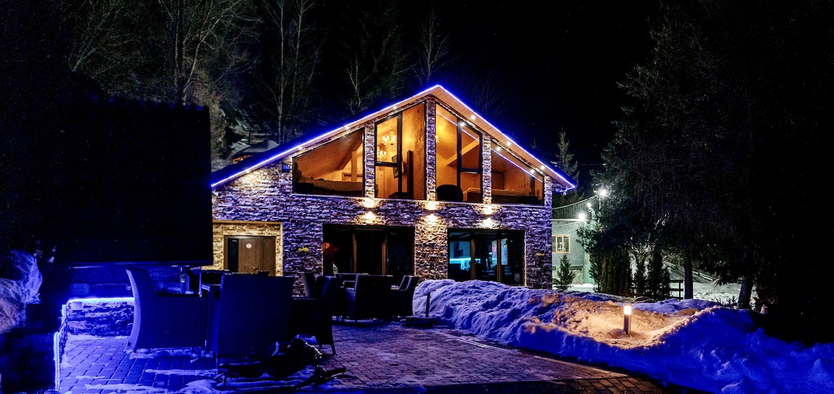 The modern villa is illuminated by soft blue lights against a dark backdrop, showcasing its stone exterior and large windows. A gathering area is visible outside, with seating arranged on a snowy ground, creating a cozy atmosphere for evening relaxation.