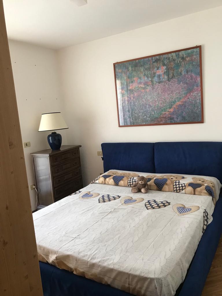 A sleeping area features a spacious bed with blue upholstery, decorated with a light blanket and a collection of soft toys. A wooden nightstand and a lamp stand alongside the bed, while a large framed artwork adorns the wall, adding a touch of color to the room.