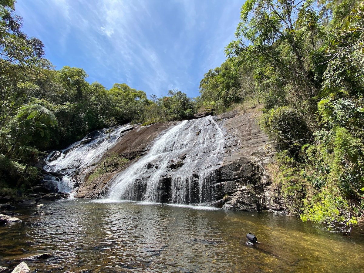 A cascading waterfall flows over a rocky surface, surrounded by dense green foliage. The tranquil pool at the base reflects the clear blue sky while the sunlight creates shimmering patterns on the water's surface.