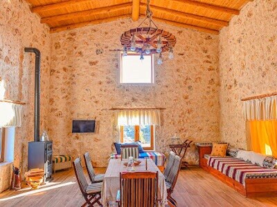 The living area features rustic stone walls and a wooden ceiling, with a long dining table surrounded by chairs. A small TV is mounted on the wall, while a cozy seating area includes colorful cushions and a futon. Natural light pours in through the windows.