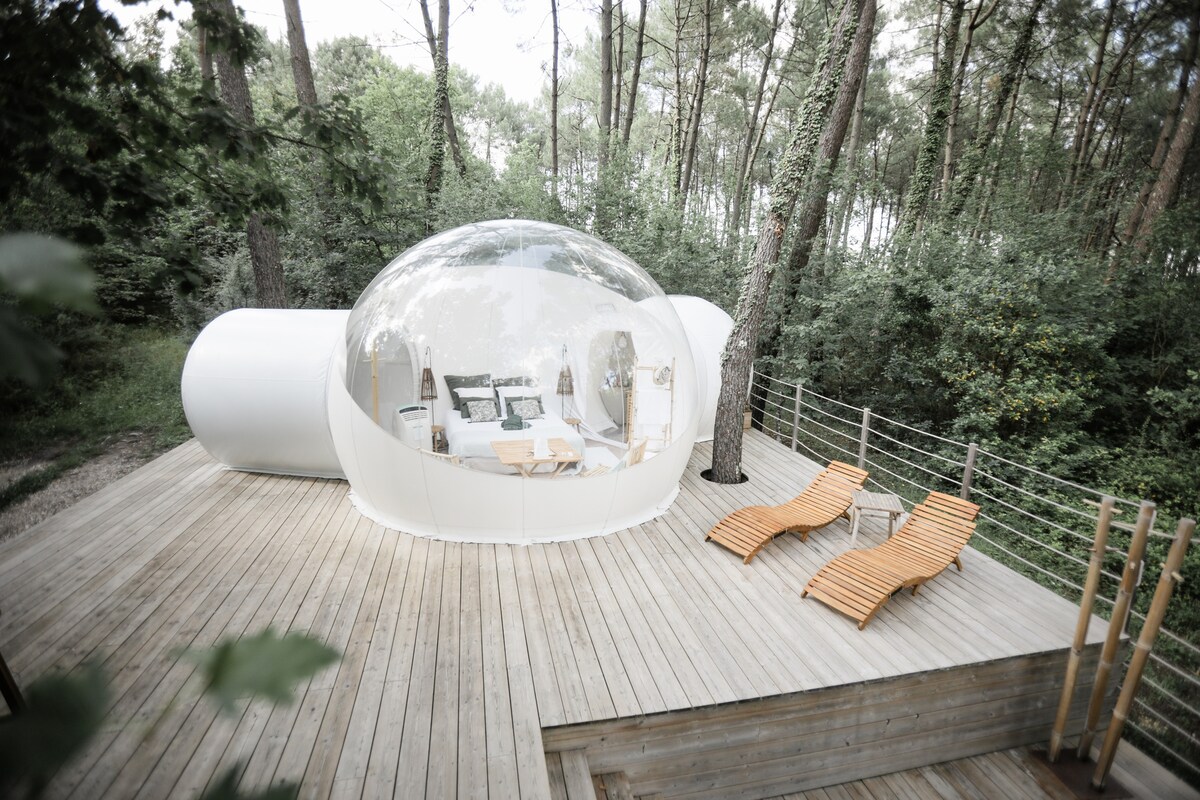 A semi-transparent bubble structure is placed on an elevated wooden terrace, surrounded by lush greenery. Two wooden sun loungers are positioned on the deck, providing a space for relaxation and enjoying the natural surroundings.