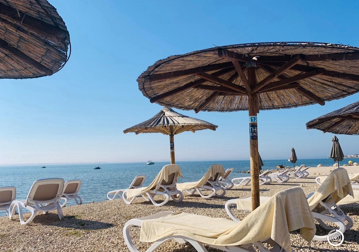A sandy beach is lined with sun loungers and thatched umbrellas providing shade. The calm sea stretches in the background, with small boats visible on the horizon. The warm, sunny setting creates an inviting spot for relaxation.