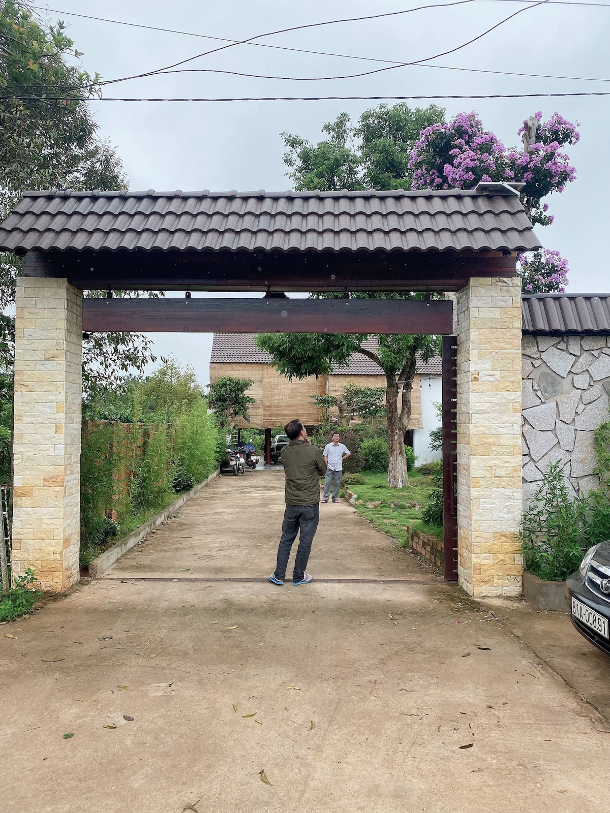 An entrance gate with a tiled roof is framed by stone columns. A wide driveway leads toward a similar structure in the background. Lush greenery surrounds the path, and cars can be seen parked along the sides.