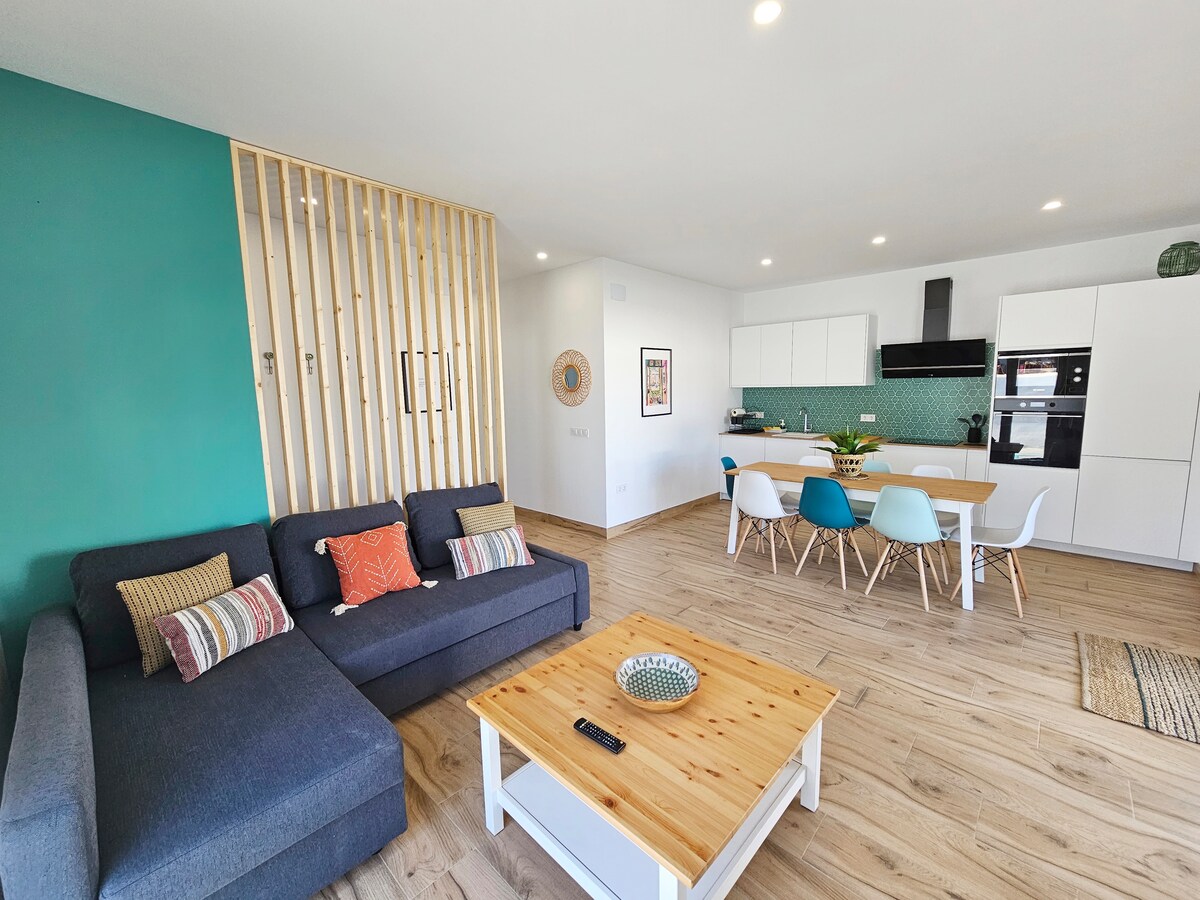 An open-plan living area is visible, featuring a modern gray sofa paired with accent pillows. A light wooden coffee table is centrally placed. The dining space includes a white table surrounded by colorful chairs, complemented by a sleek kitchen with cabinetry and appliances.