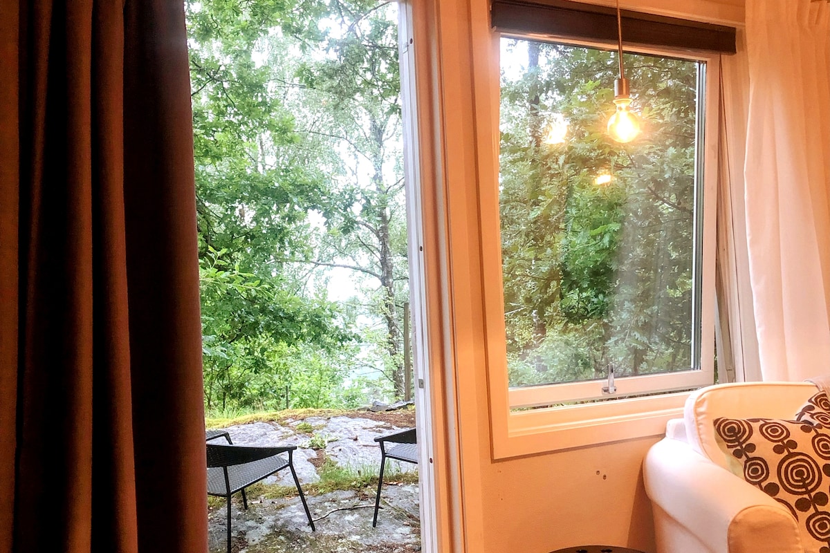 A window showcases a view of a serene outdoor area, framed by lush greenery. Two outdoor chairs are positioned on a natural stone surface, inviting relaxation. Soft lighting is provided by a pendant light hanging from the ceiling.