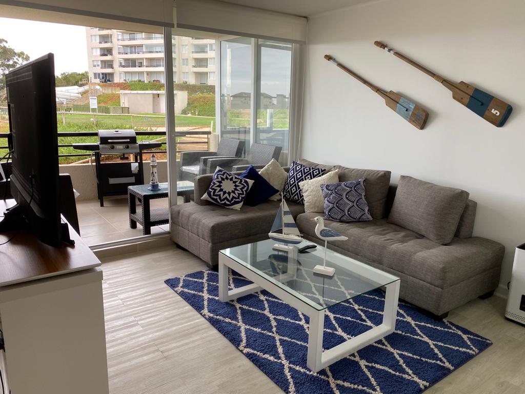 A cozy living area features a comfortable gray sectional sofa adorned with decorative pillows. A glass coffee table sits atop a blue patterned rug. A large window allows natural light to fill the space, with decorative acoustic guitars mounted on the wall above.