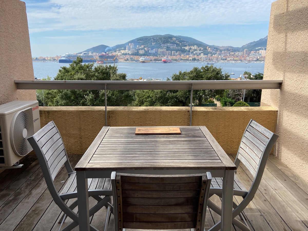 A wooden dining table with four chairs is positioned on a terrace, offering a view of the distant hills and the sea. The scene includes a calm sky with scattered clouds, enhancing the outdoor dining experience.