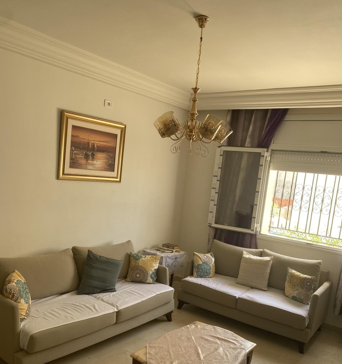 A cozy living area features two light-colored sofas adorned with decorative cushions in soft hues. A small coffee table is positioned between them. Natural light enters through a window, which is framed by sheer curtains, while a chandelier adds a touch of elegance to the space.