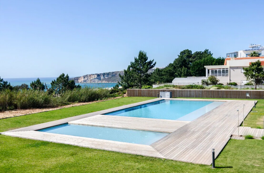 The image displays a spacious outdoor area featuring two rectangular swimming pools framed by a wooden deck. Lush green landscaping surrounds the pools, with views of the ocean and cliffs in the background, creating a serene environment.