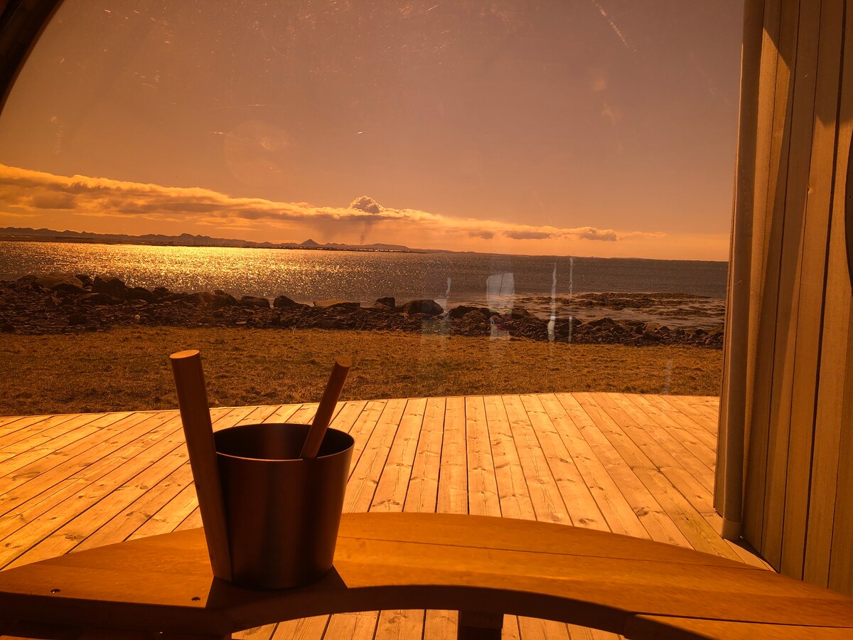 A serene view is captured through a curved glass wall, highlighting the sunlit sea and distant shoreline. A wooden table is positioned in the foreground, featuring a bowl, adding a touch of elegance to the tranquil setting.