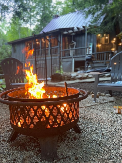 A firepit is positioned in a gravel area, surrounded by dark Adirondack chairs. A flickering flame creates a warm glow, drawing attention to the treehouse visible in the background. Soft lighting adorns the porch area, enhancing the tranquil outdoor setting.