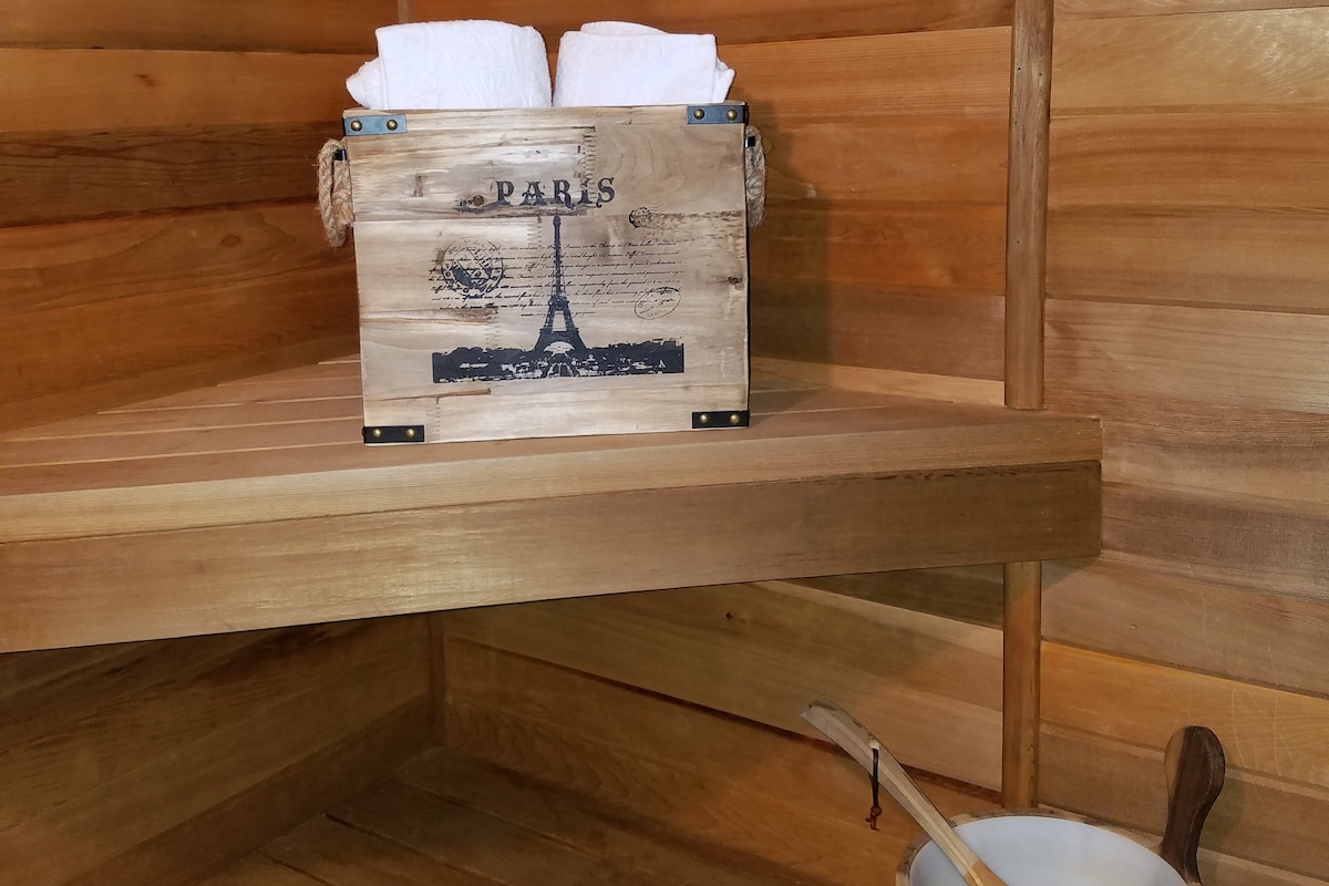 The wooden sauna interior is illuminated softly, featuring smooth, warm wooden panels throughout. A decorative box labeled 'Paris' holds neatly stacked white towels, placed on a wooden shelf. A wooden ladle and a small bucket are positioned nearby, contributing to the inviting ambiance.