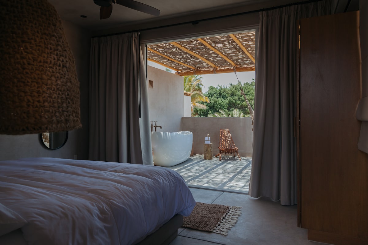A spacious bedroom features a plush bed with crisp linens. Natural light floods the room, illuminating a private terrace with a freestanding bathtub and a cozy chair. Soft drapes frame the entrance, enhancing the connection to the outside greenery.