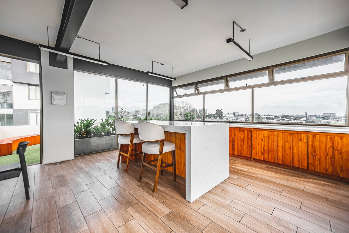 A bright and airy dining area is highlighted by large windows, offering panoramic views. Two modern bar stools are positioned near a minimalist white island countertop. Warm wooden paneling surrounds the space, complementing the light wooden flooring and providing an open and inviting atmosphere.