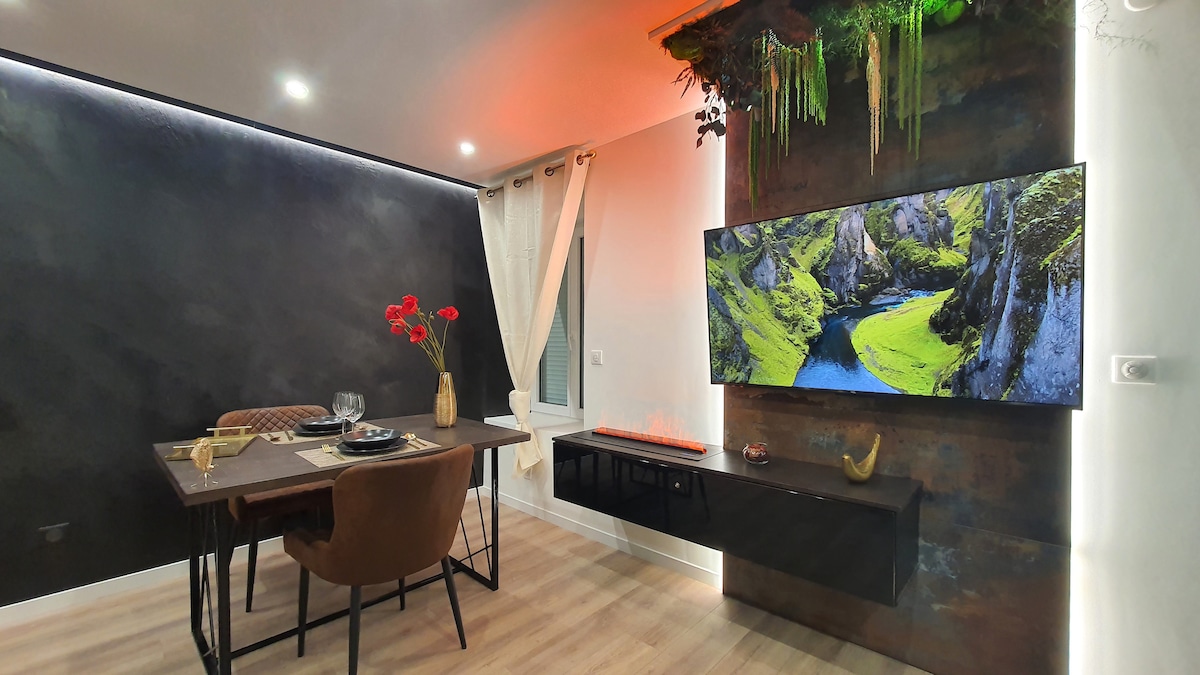 A modern living space features a wall-mounted television displaying a serene landscape. A dining table is set for two, accompanied by stylish chairs. Soft lighting highlights the earthy decor, including natural elements cascading from the wall, while large windows allow for natural light.