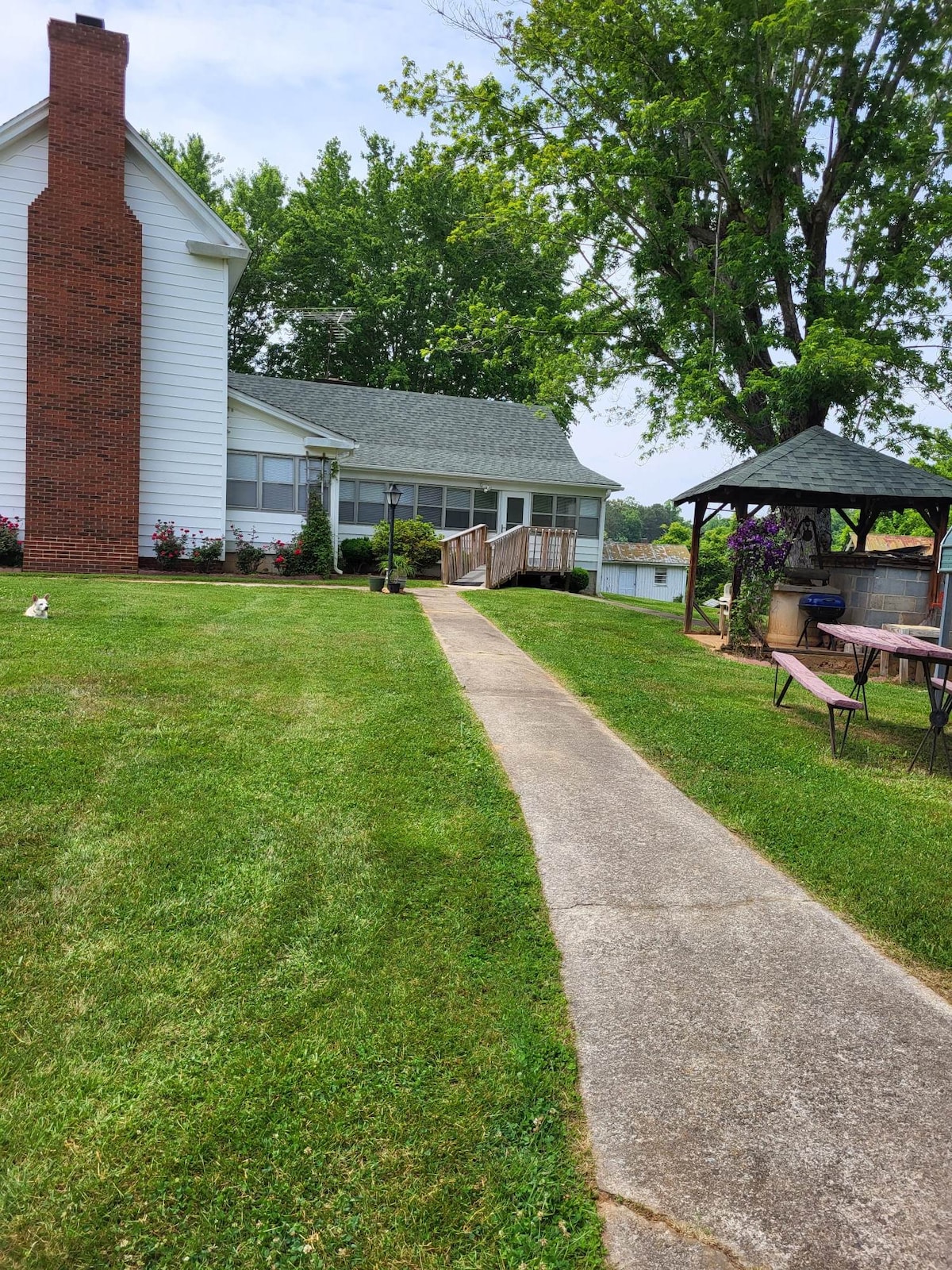 A well-maintained pathway leads towards a charming farmhouse surrounded by lush green grass and colorful flower beds. A gazebo can be seen in the distance, offering a shaded area for relaxation. The entrance features a gentle ramp for accessibility.
