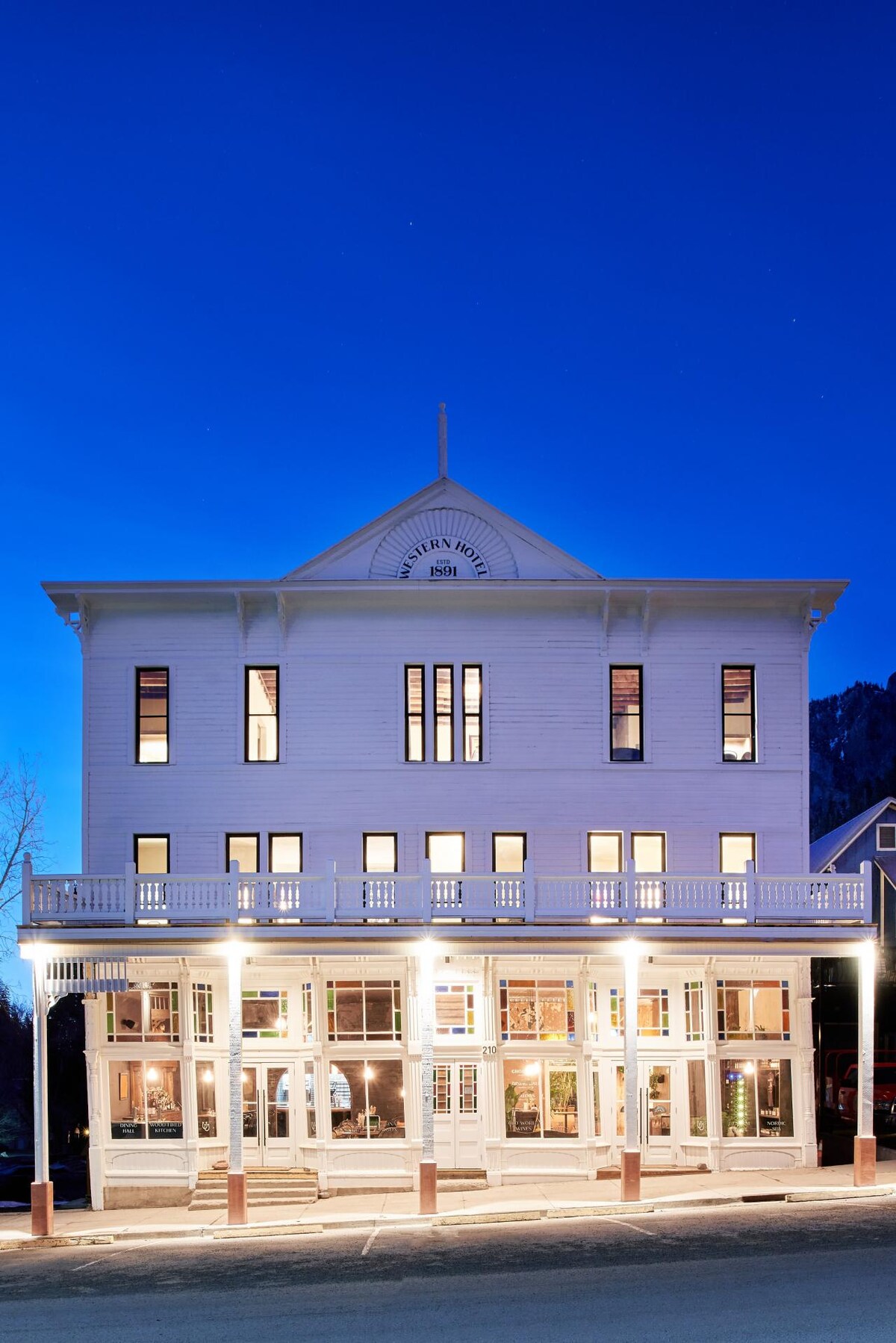 The exterior of The Western Hotel & Spa is depicted at twilight, showcasing its two-story white structure with elegant architectural details. Large windows are illuminated, revealing the inviting atmosphere. Outdoor seating is present in front, complemented by a well-lit walkway that enhances the building's historic charm.