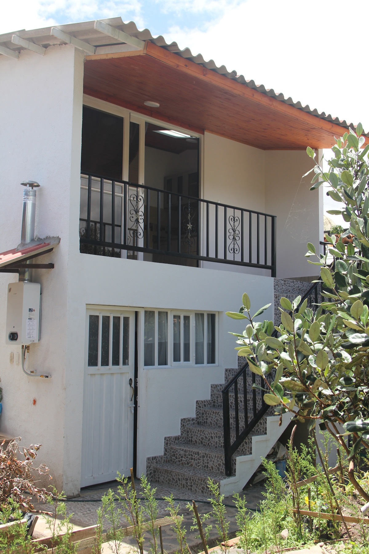 A two-story house is presented, featuring a wooden overhang above a balcony with decorative iron railings. The entrance is marked by a white door, leading to a ground-level space with a textured stone staircase. Lush greenery surrounds the exterior, enhancing the natural setting.