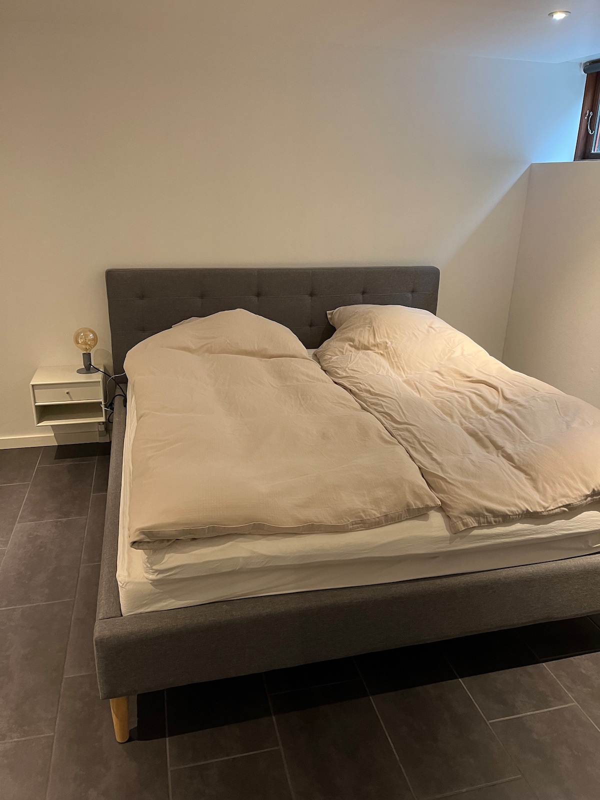 A spacious sleeping area features a neatly made bed with a light-colored duvet, complemented by two separate comforters. A nightstand is positioned next to the bed, with a small lamp displayed on top. Natural light enters through a nearby window, enhancing the room's brightness.