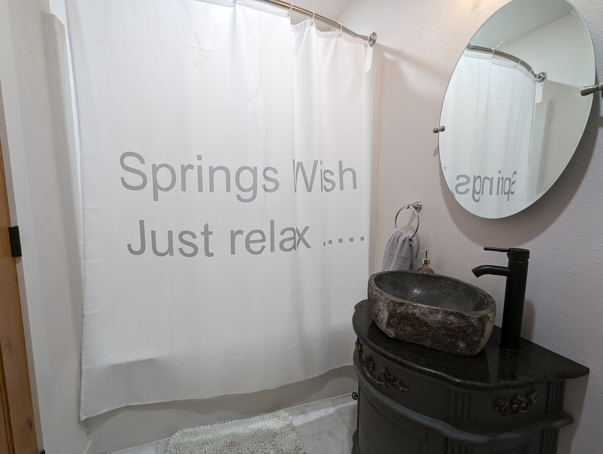 A bathroom is featured with a calming shower curtain displaying 'Springs Wish Just relax ....'. A stone vessel sink rests on a dark vanity, complemented by a round mirror. Soft bath accessories enhance the serene atmosphere.