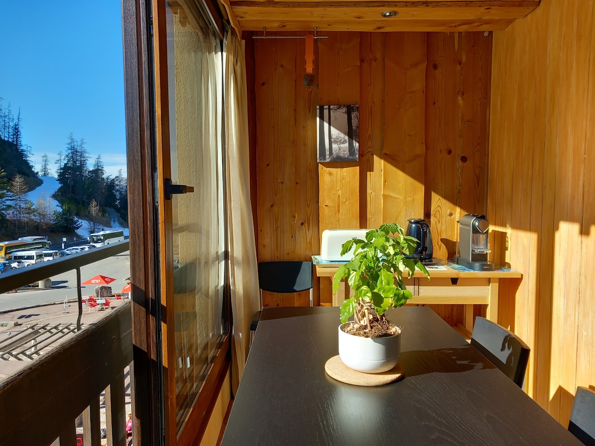 A sunlit loggia features warm wooden walls, with a small table and black chairs positioned beside a potted plant. A Nespresso coffee machine is visible on a counter, providing a cozy space to enjoy a morning beverage.