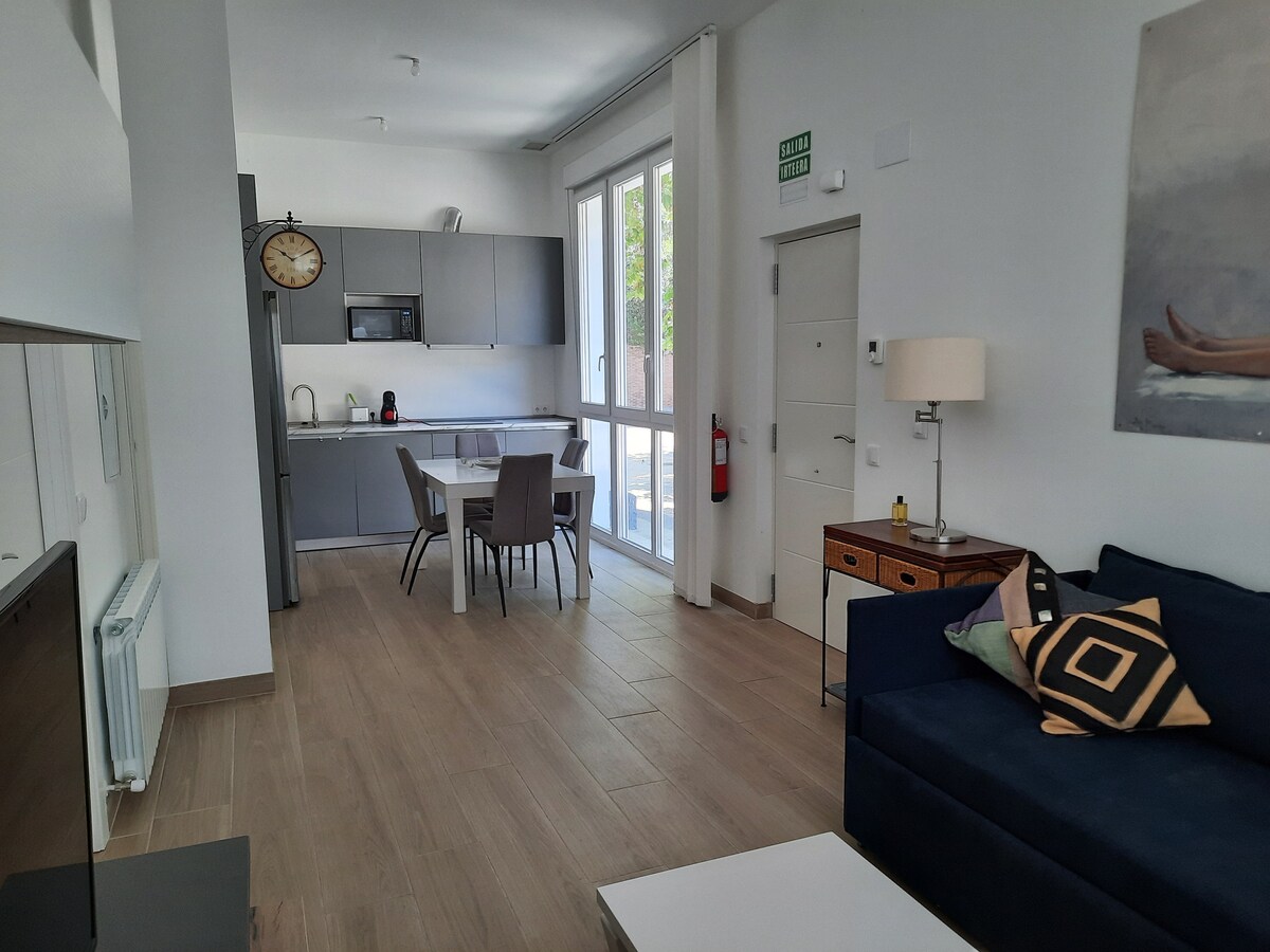An open-plan living area features a comfortable navy sofa and a modern dining table with chairs. The kitchen area is equipped with contemporary appliances. Large windows allow natural light to fill the space, and light wooden flooring adds warmth to the design.