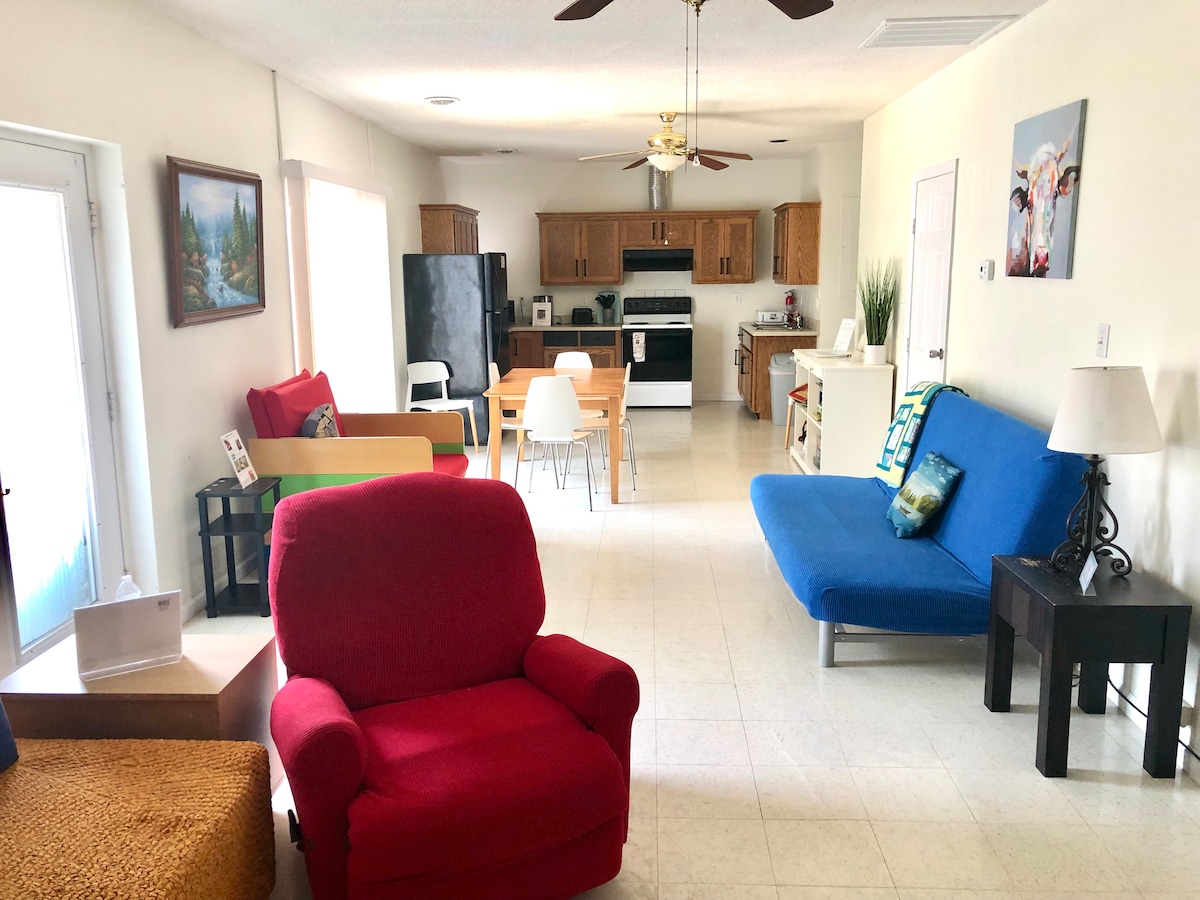 An open living space features a red armchair and a blue futon across from a dining area with a wooden table and chairs. Natural light fills the room through large windows, illuminating the kitchen equipped with wooden cabinets and modern appliances.