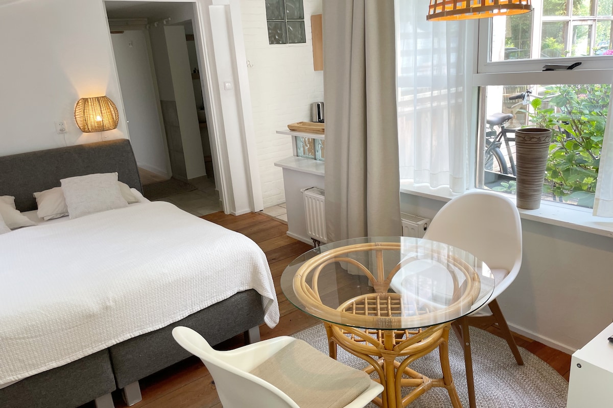 A well-lit room features a spacious bed with light linens and a round glass table surrounded by two white chairs. A grey curtain partially covers the window, which offers a view of greenery outside. A cozy atmosphere is created by soft lighting and warm wooden accents.