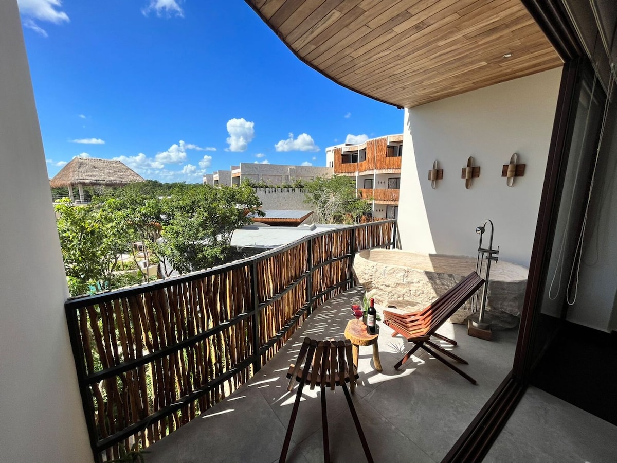 A spacious terrace is featured, offering a view of lush greenery and neighboring structures. Two wooden loungers are positioned for relaxation, accompanied by a small table. Natural light is welcomed through large glass doors, emphasizing the connection to the outdoor environment.
