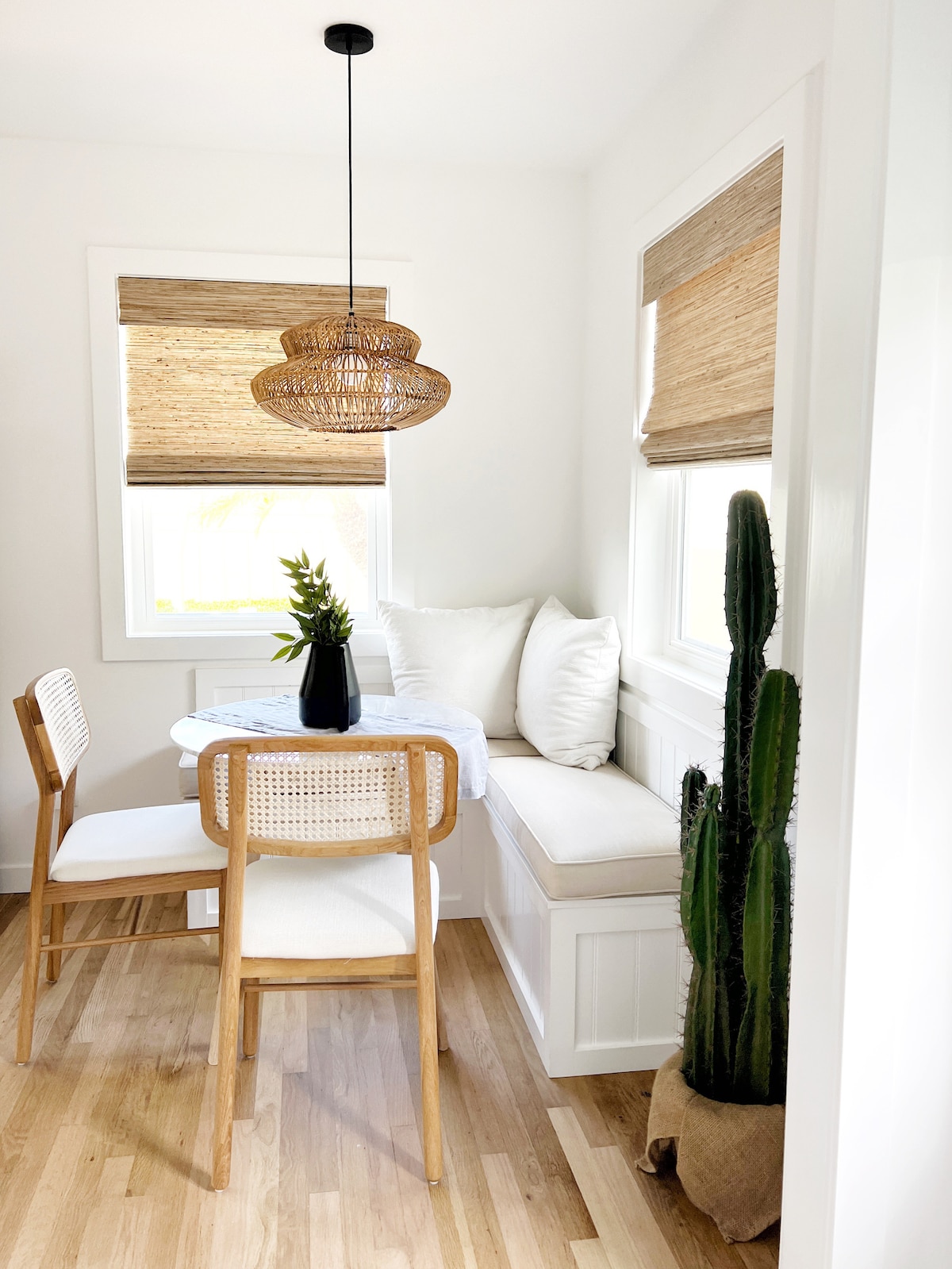 A dining nook features a round table surrounded by two chairs, with a built-in bench adorned with white cushions. Natural light filters through woven shades on the windows, while a blooming plant and a tall cactus add greenery to the bright, clean space.