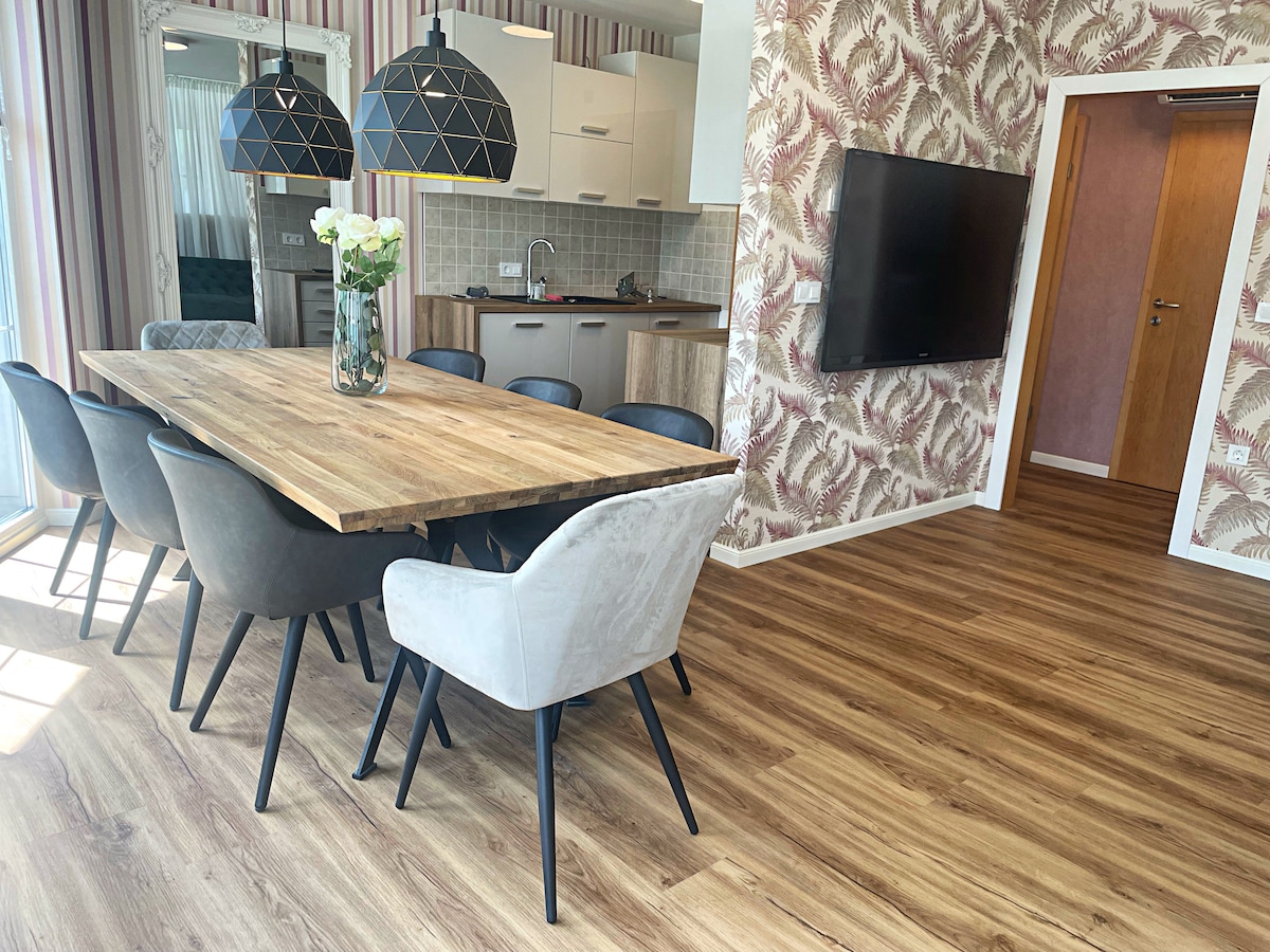 A spacious dining area is highlighted by a large wooden table surrounded by six chairs. Two pendant lights hang above, while a floral centerpiece adds a touch of elegance. A television is mounted on the wall nearby, complementing the modern aesthetic of the room.