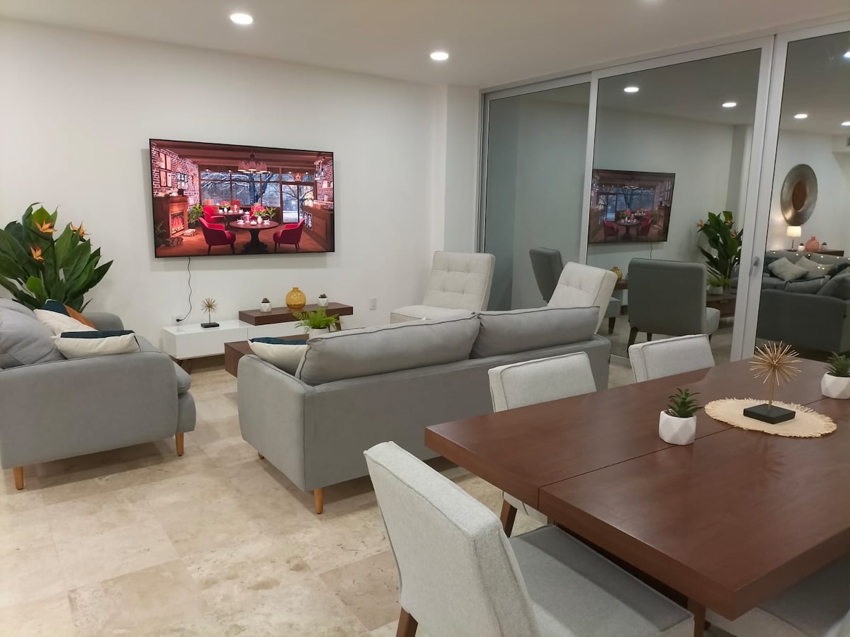 A well-furnished living area features two grey sofas facing a large screen television. A wooden dining table with light-colored chairs occupies the space, and decorative plants add greenery to the neutral palette. Natural light enters through expansive glass doors.