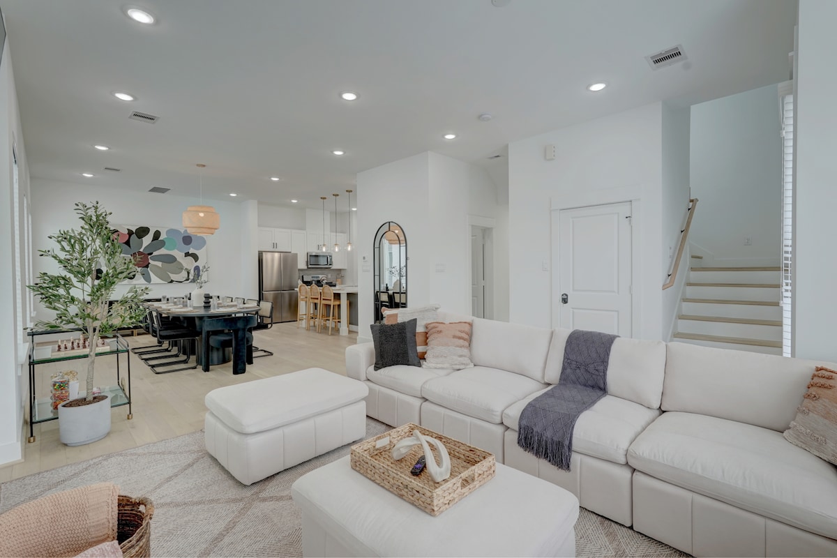 A spacious living room showcases a light-colored sectional sofa, paired with a neutral coffee table. A plant adds a touch of greenery, while recessed lighting illuminates the area. In the background, an open dining space with a large table and modern kitchen appliances is visible.