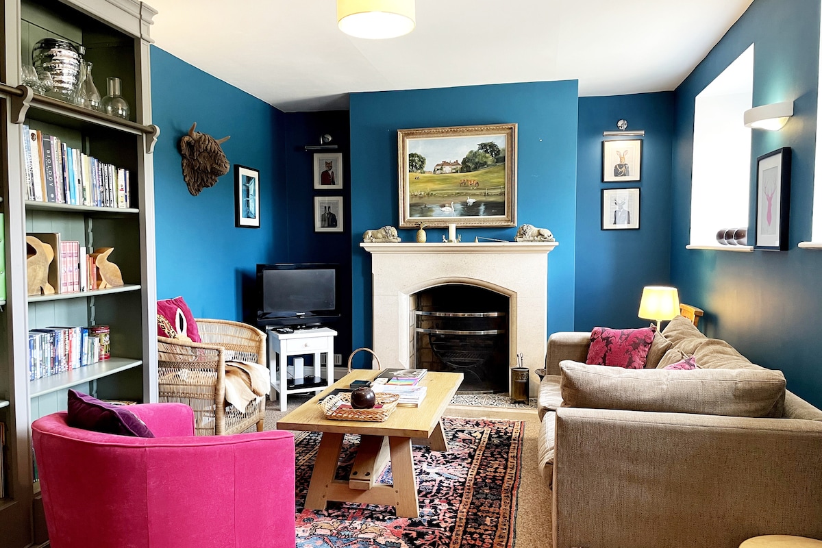 A snug sitting room features a striking blue wall with a sandstone open fireplace as the focal point. Comfortable seating includes a beige sofa and two accent chairs. A coffee table sits at the center, surrounded by a vintage rug. Natural light filters through nearby windows.
