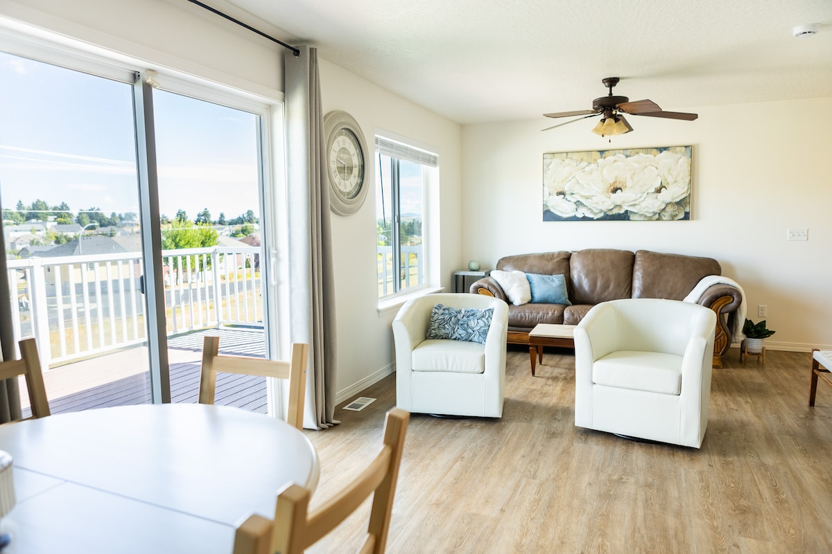 The living area features a comfortable brown sofa paired with two white armchairs, offering a welcoming space. Large sliding glass doors provide natural light and open to a scenic view, complemented by a soft ceiling fan and decorative wall art.