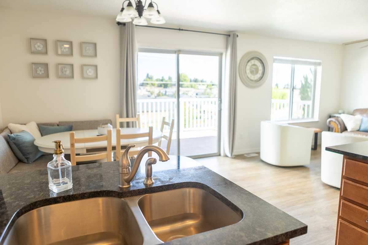 A kitchen area is visible, featuring a double sink and a modern faucet. In the background, a dining table with chairs is seen alongside a cozy seating arrangement. Large windows allow natural light to flood the space, with access to a balcony enhancing the open feel.