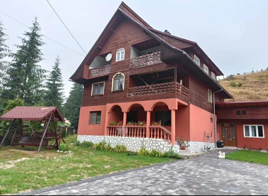 A three-story wooden house with a red exterior is surrounded by lush greenery. A swing set is positioned in the yard, alongside a stone pathway leading to the entrance. Balconies are visible on the upper floor, enhancing the charming exterior.