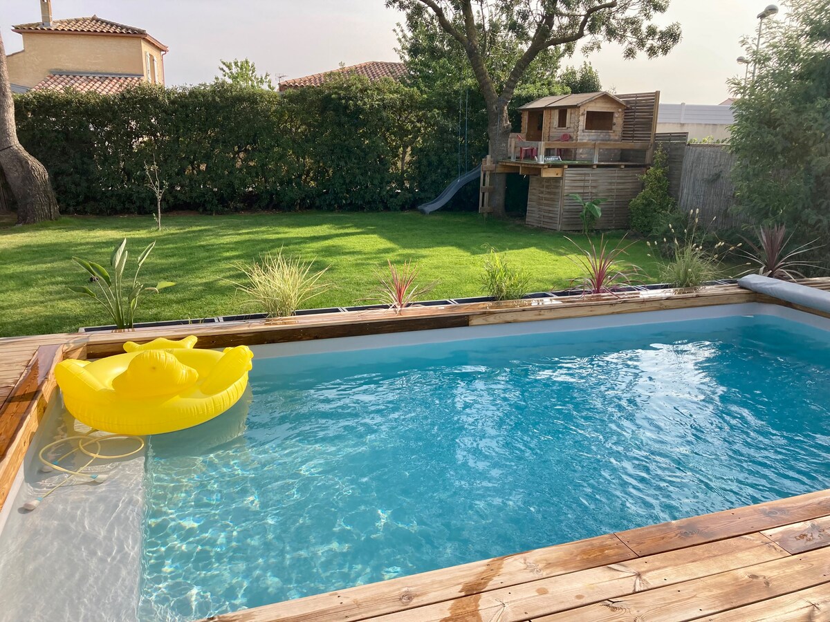 A serene outdoor space features a sparkling pool surrounded by a wooden deck. A vibrant yellow float is placed in the water. Lush green grass and decorative plants adorn the area, while a wooden playhouse and slide can be seen in the background, enhancing the family-friendly atmosphere.