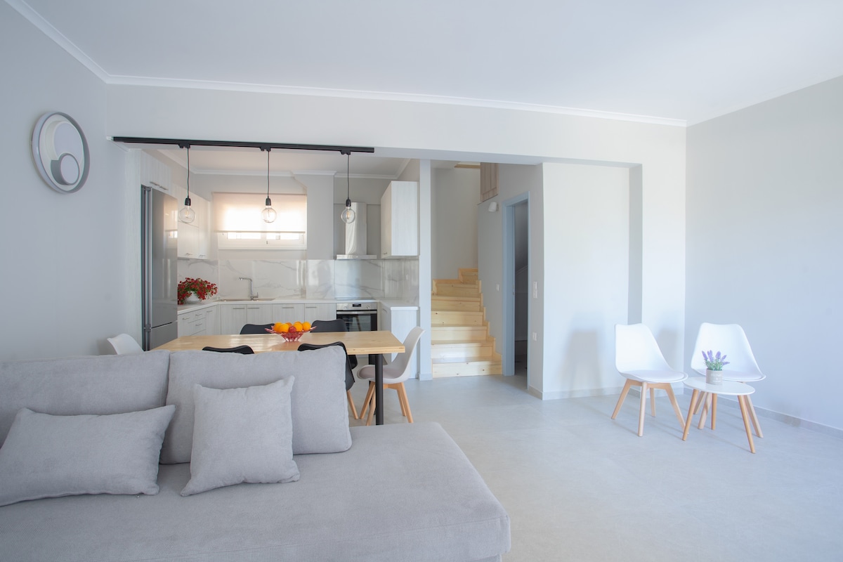 A modern living area features a light gray sofa, facing a dining table set for four. A sleek kitchen is visible in the background, equipped with white cabinetry. Natural light floods the space through large windows, enhancing the clean, minimalist aesthetic.