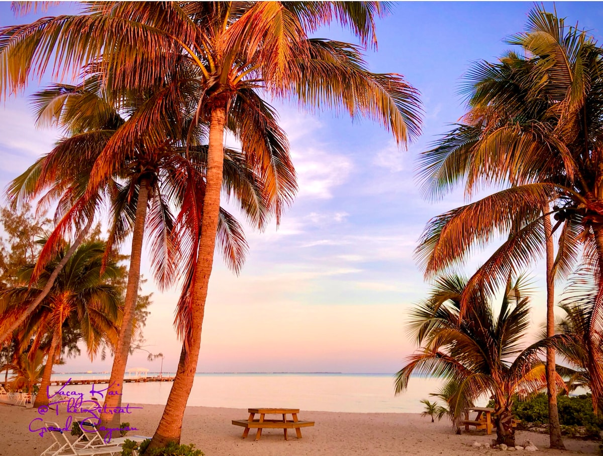 Tall palm trees frame a sandy beach at sunset, with shades of pink and orange in the sky. A wooden picnic table is visible beneath the trees, providing a shaded area for relaxation near the aquamarine water.