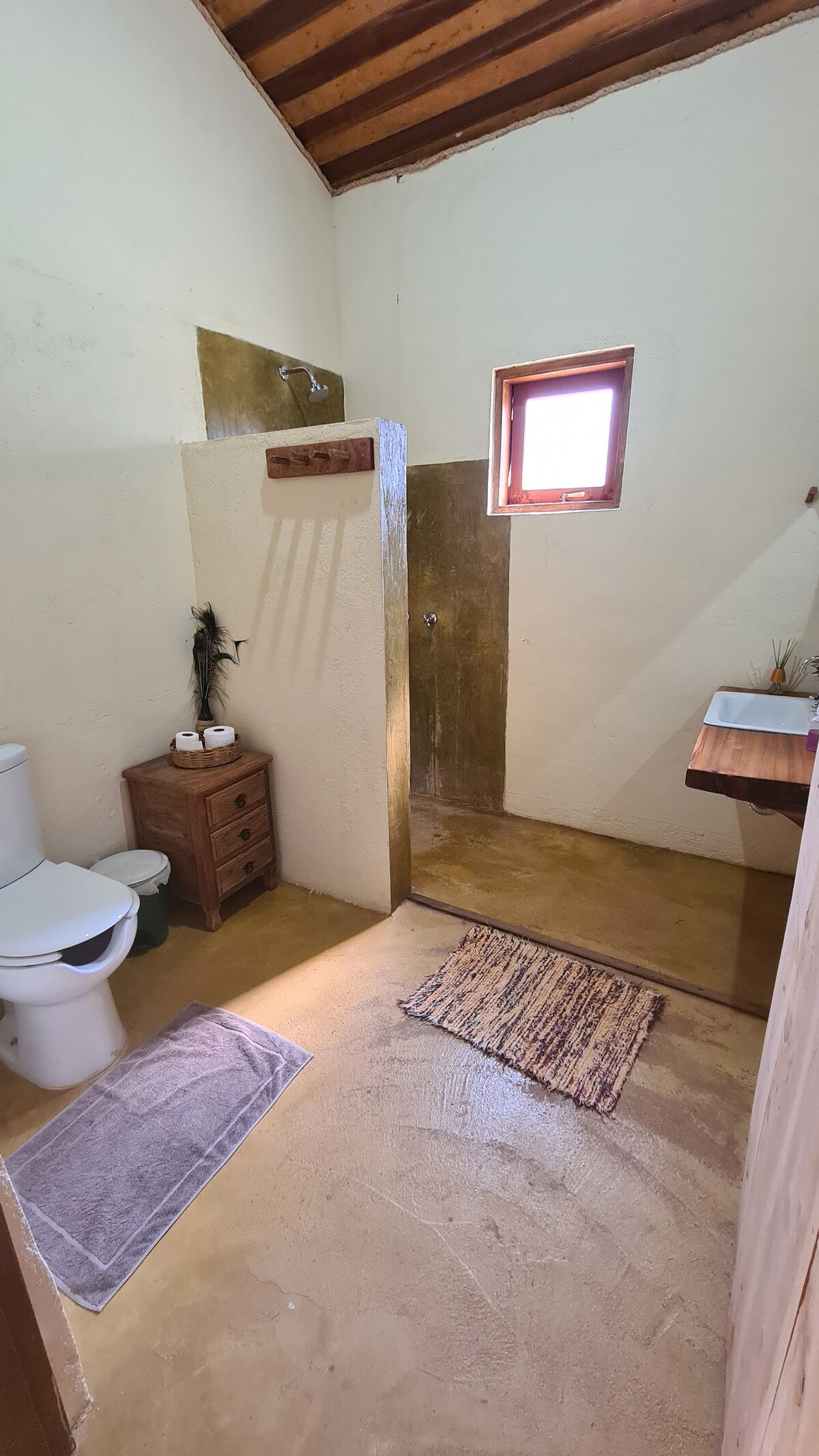 The bathroom features a minimalist design with beige walls and a tiled shower area. A wooden shelf above the sink offers toiletries, while natural light filters in through a small window. A soft rug lies on the floor, adding comfort to the space.