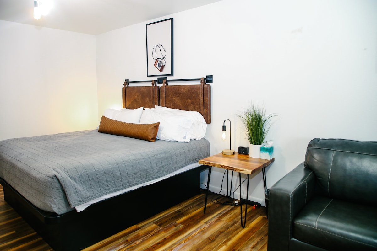 A neutral-colored room features a spacious queen bed with a stylish headboard and crisp linens. A small bedside table with a lamp sits next to the bed. A comfortable chair complements the modern wooden flooring, creating a cozy seating area.
