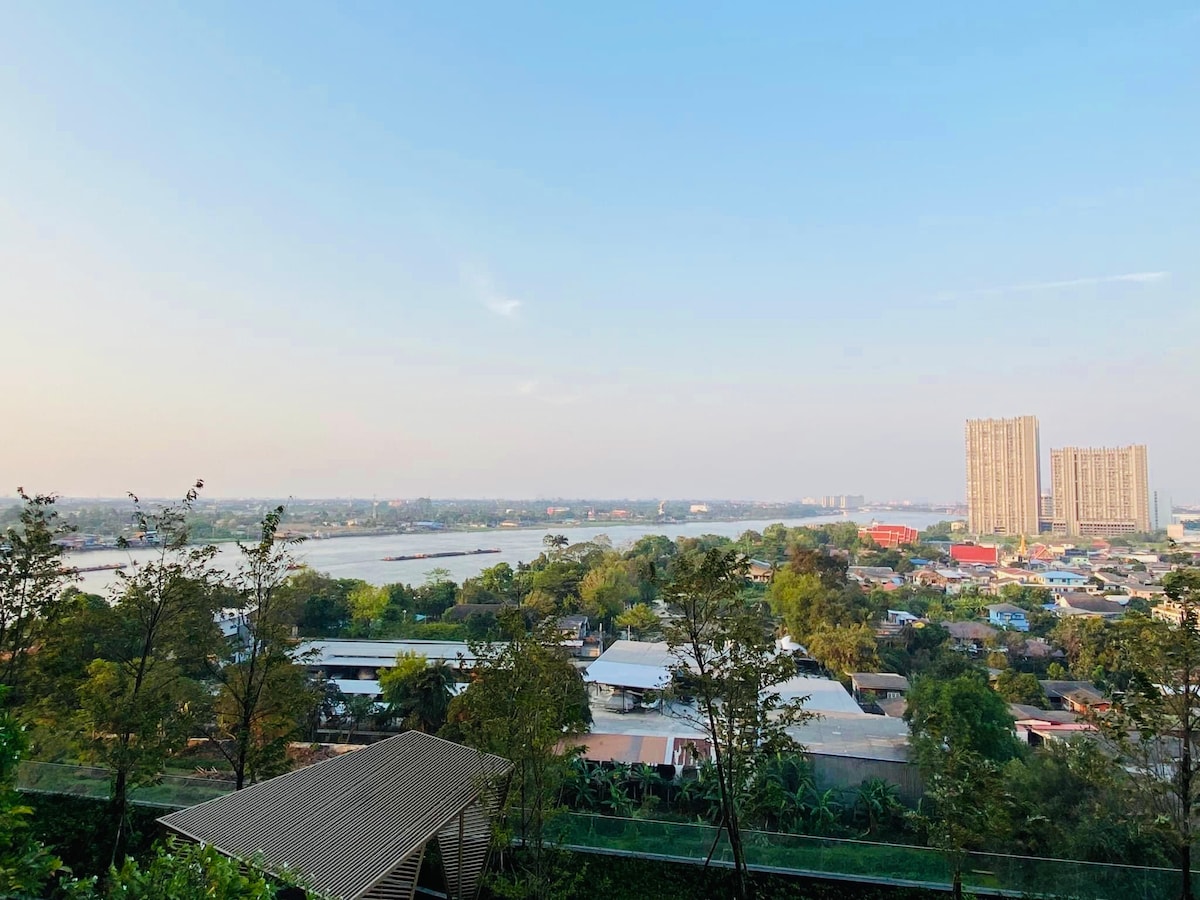 A scenic view is captured from the balcony, showcasing the river alongside a lush landscape. Urban buildings rise in the distance, while greenery and residential areas create a harmonious balance. The sky is clear, hinting at a calm atmosphere.